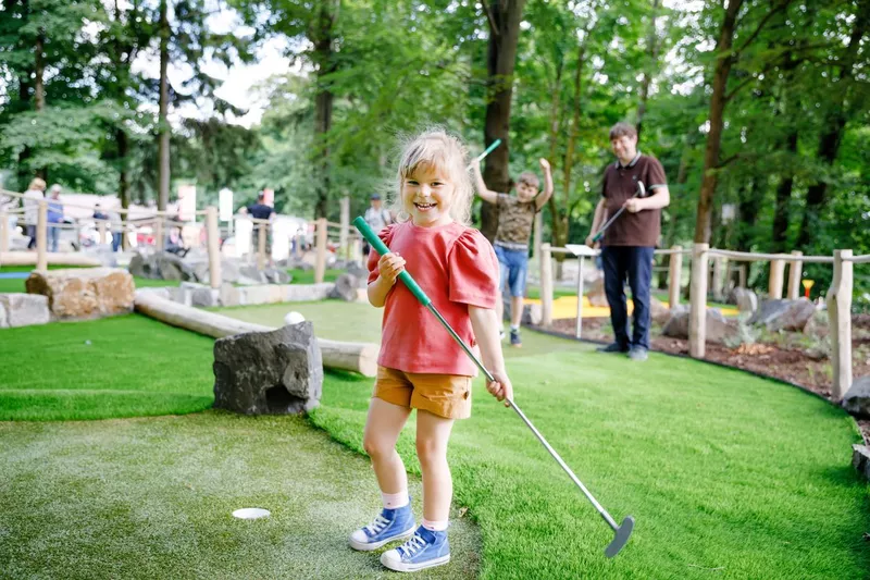 Cute preschool girl playing mini golf with family. Happy toddler child having fun with outdoor activity. Summer sport for children and adults, outdoors. Family vacations or resort.